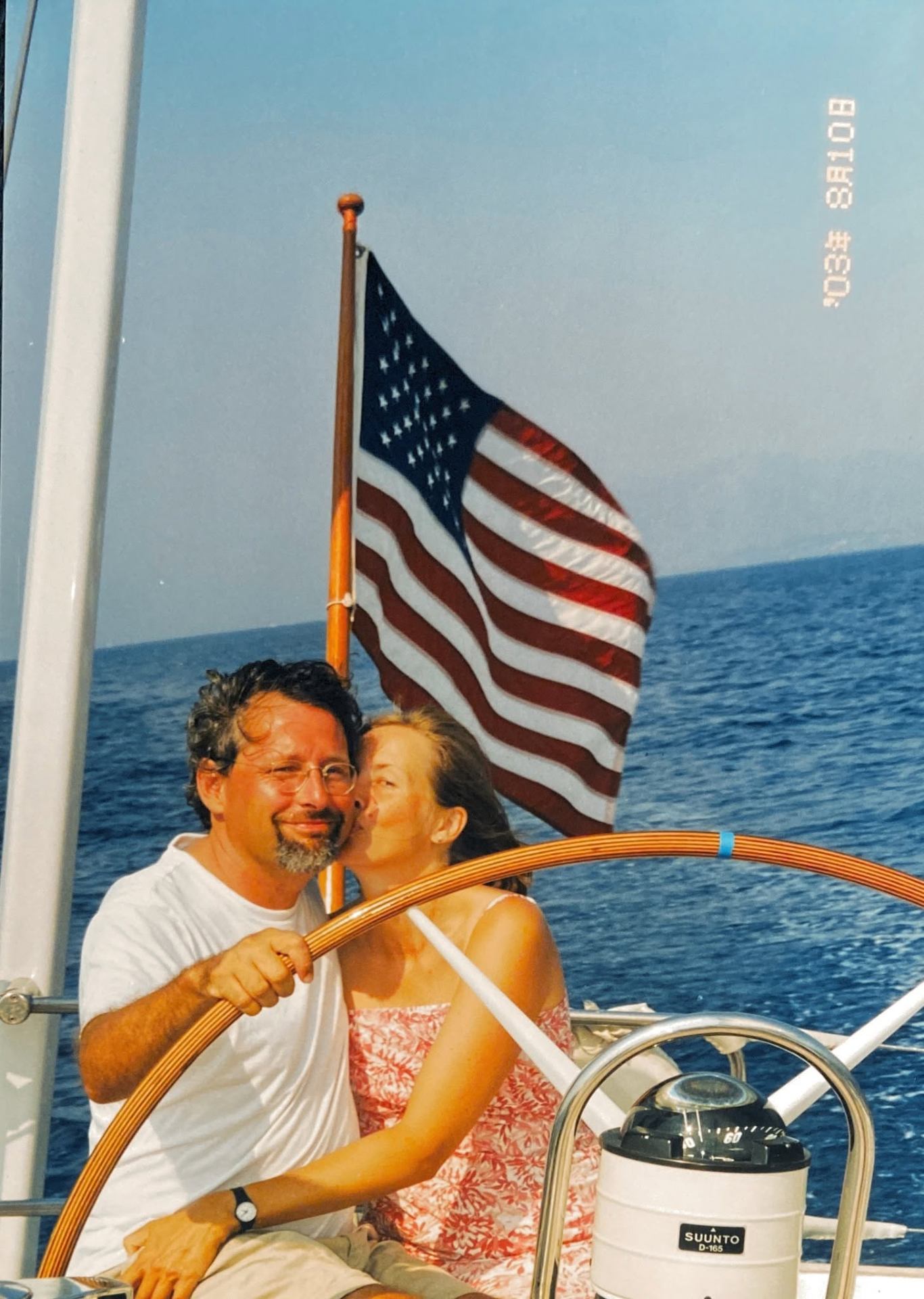 Pierre at the helm of a sailboat with his wife, an American flag flying behind them.