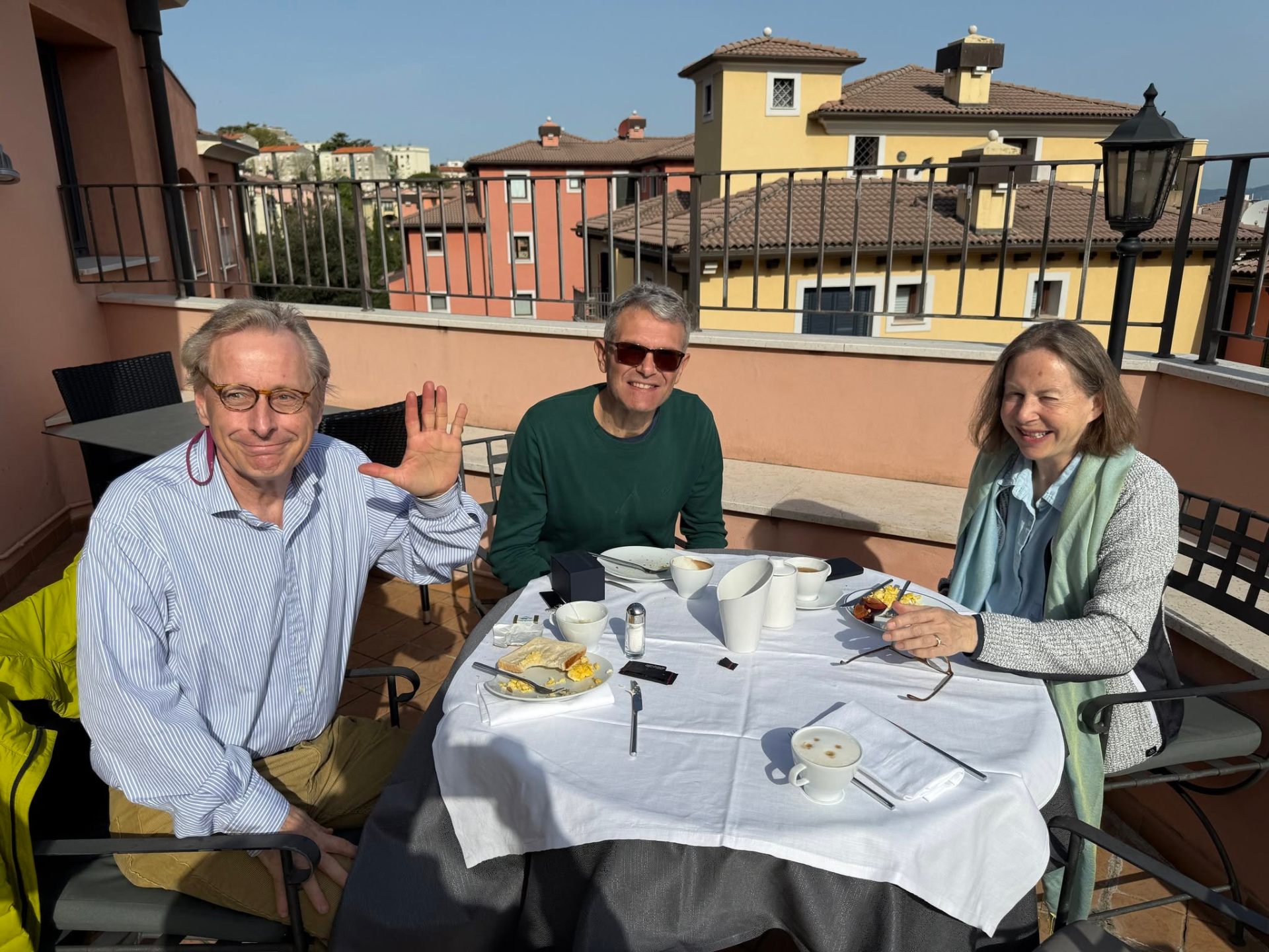 Pierre waving at a sunny rooftop breakfast in Italy with two friends.